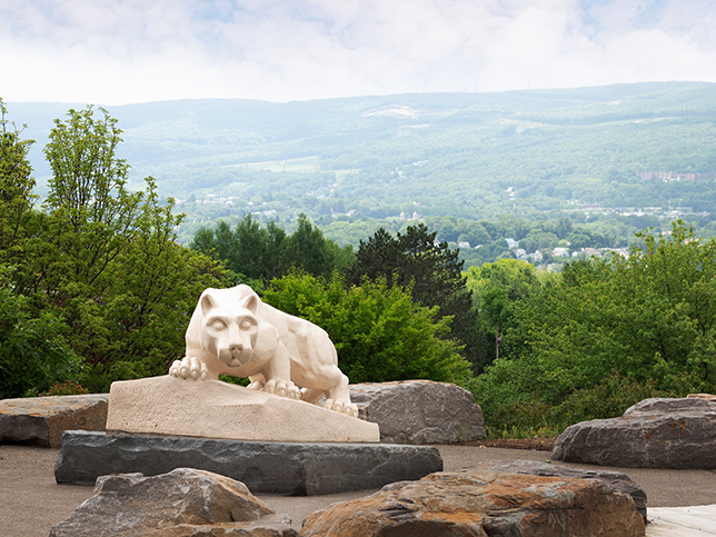 There are boulders in the foreground, the Nittany Lion Shrine in the mid-ground, and lush trees, blue skies, and mountains in the background.